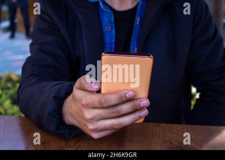 Jeune homme tenant et utilisant un smartphone tout en étant assis dans un café. Banque D'Images
