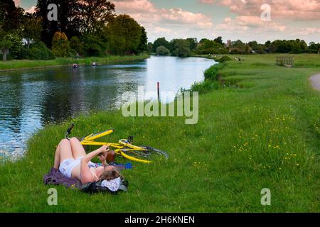 Jeune fille se détendant seule sur des prairies d'herbe regardant son téléphone avec cycle jaune sur la rive de la rivière Cam près de Cambridge Angleterre Banque D'Images