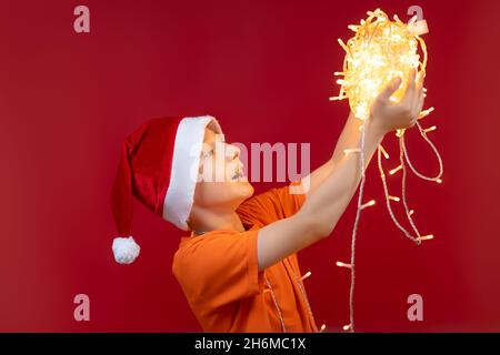 Un garçon gai dans un chapeau de Père Noël sur les bras étendus tient une pelote de guirlandes lumineuses devant lui Banque D'Images
