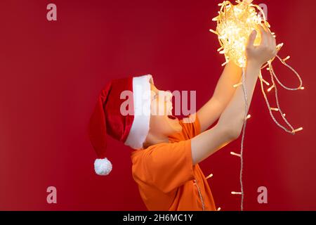 Un garçon gai dans un chapeau de Père Noël sur les bras étendus tient une pelote de guirlandes lumineuses devant lui Banque D'Images