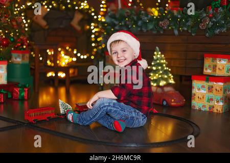 Un petit garçon gai dans le chapeau du Père Noël joue avec la locomotive à la maison tout en étant assis près de l'arbre de Noël. Banque D'Images