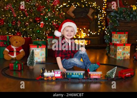 Le petit garçon heureux dans le chapeau du Père Noël joue avec la locomotive à la maison tout en étant assis près de l'arbre de Noël. Banque D'Images