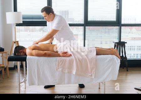 Photo de côté d'un masseur professionnel masculin avec de fortes mains tatouées massant le dos et l'épaule d'un sportif musclé allongé sur l'estomac Banque D'Images