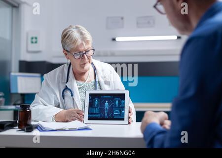 Médecin analysant le comprimé avec l'illustration de l'analyse du corps humain avec l'homme ancien.Un spécialiste de la santé explique le diagnostic au patient malade tout en tenant l'appareil avec une anatomie virtuelle et une expertise Banque D'Images