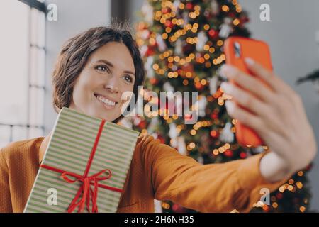 Belle brune femelle prenant selfie avec boîte cadeau de Noël près de l'arbre de Noël décoré à la maison Banque D'Images