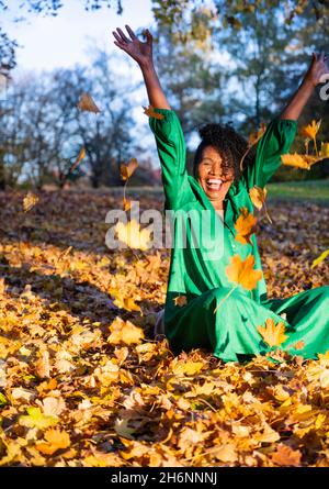 Portrait d'une femme rieuse à la peau sombre avec des boucles dans une robe verte, assis et jetant des feuilles d'automne, prise en plein air en automne, Allemagne Banque D'Images