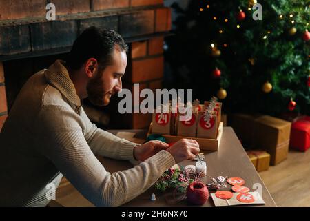 Beau jeune homme écrivant le numéro sur le sac rouge faisant des sacs en papier de papier kraft pour le calendrier de l'Avent la veille de Noël assis près d'une cheminée et d'un arbre. Banque D'Images