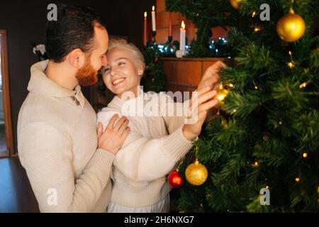 Photo moyenne de charmant jeune couple en amour décorant l'arbre de Noël avec des boules brillantes près de la cheminée dans le salon sombre confortable. Banque D'Images
