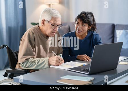 Homme senior assis à la table à la maison et remplissant des documents avec l'aide de bénévoles Banque D'Images