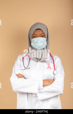 Portrait d'une femme afro-américaine portant un masque chirurgical, une écharpe et un manteau blanc posant avec ses bras croisés. Banque D'Images