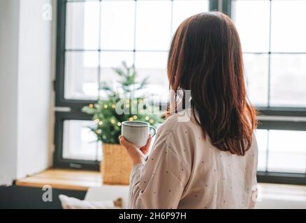 Jeune adulte quarante ans femme avec cheveux foncés dans le confortable pyjama avec tasse de thé dans le salon avec arbre de Noël, les gens de derrière Banque D'Images