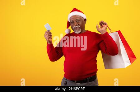 Ventes et achats de Noël.Homme noir senior avec sacs cabas tenant une carte de crédit, portant chapeau de père Noël Banque D'Images