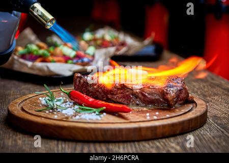 Mise au point douce.Entrecote boeuf steak grillé viande avec flammes de feu sur bois de coupe avec branche de romarin, poivre et sel.Cuisine du chef d Banque D'Images