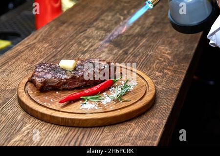 Mise au point douce.Entrecote boeuf steak grillé viande avec flammes de feu sur bois de coupe avec branche de romarin, poivre et sel.Cuisine du chef d Banque D'Images