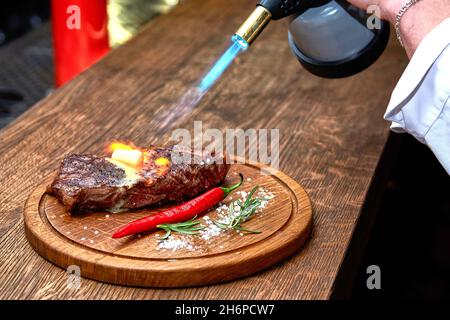 Mise au point douce.Entrecote boeuf steak grillé viande avec flammes de feu sur bois de coupe avec branche de romarin, poivre et sel.Cuisine du chef d Banque D'Images