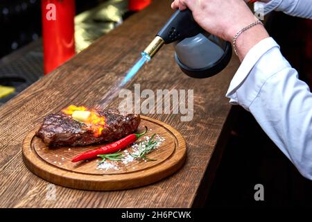 Mise au point douce.Entrecote boeuf steak grillé viande avec flammes de feu sur bois de coupe avec branche de romarin, poivre et sel.Cuisine du chef d Banque D'Images