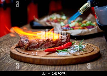 Mise au point douce.Entrecote boeuf steak grillé viande avec flammes de feu sur bois de coupe avec branche de romarin, poivre et sel.Cuisine du chef d Banque D'Images