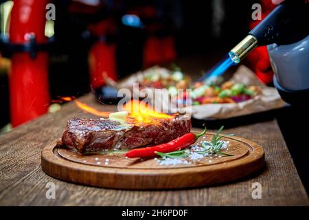 Mise au point douce.Entrecote boeuf steak grillé viande avec flammes de feu sur bois de coupe avec branche de romarin, poivre et sel.Cuisine du chef d Banque D'Images