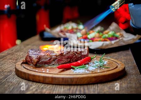 Mise au point douce.Entrecote boeuf steak grillé viande avec flammes de feu sur bois de coupe avec branche de romarin, poivre et sel.Cuisine du chef d Banque D'Images