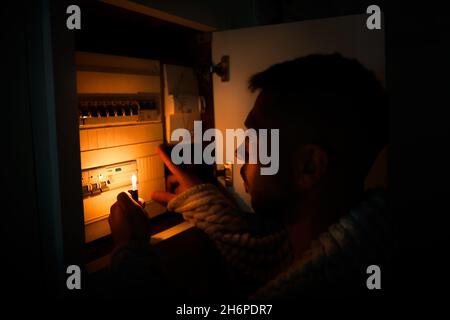 Homme avec un briquet dans l'obscurité totale étudiant la boîte à fusibles ou le tableau électrique à la maison pendant la coupure de courant.Rideaux occultants, pas d'électricité Banque D'Images