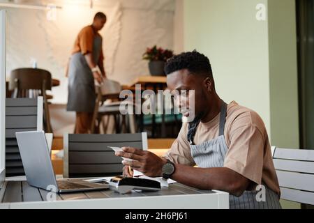 Un jeune homme portant un tablier assis à une table devant un ordinateur portable dans un café moderne prenant des commandes en ligne de clients pour la livraison Banque D'Images