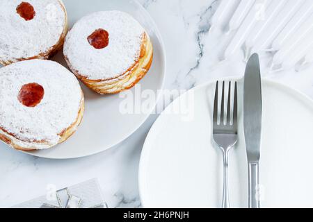 Table pour les vacances juives de Hanukkah.Assiette et couverts sur fond blanc.Bougies, cadeaux et dessert beignet sufganiyot. Banque D'Images