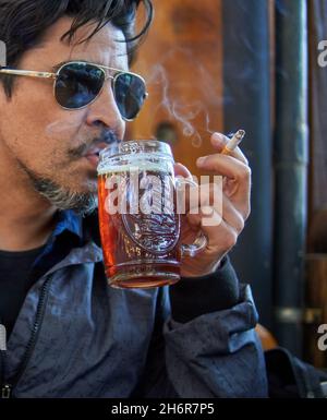 brunette latin homme en profil avec des lunettes de soleil boire une pinte de bière artisanale d'ambre et fumer une cigarette en vacances dans un bar.Verticale Banque D'Images
