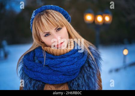 Portrait d'extérieur de jolie jeune femme aux lèvres rouges portant des vêtements chauds regardant l'appareil photo avec un petit sourire.Fille de Noël.Belle femme d'hiver dans le parc le soir.Copier l'espace Banque D'Images