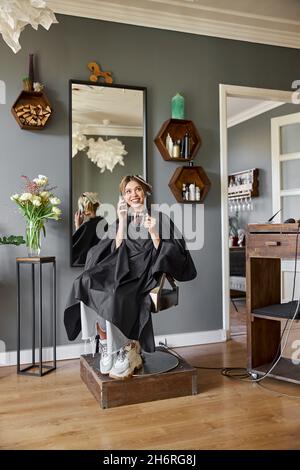 Bonne blonde la femme caucasienne utilise un smartphone tout en étant assise sur une chaise et en attendant dans le salon de coiffure Banque D'Images