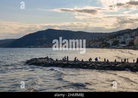 Vue panoramique de la baie avec silhouettes de personnes sur le brise-lames rocheux et la côte au coucher du soleil, Alassio, Savona, Ligurie, Italie Banque D'Images