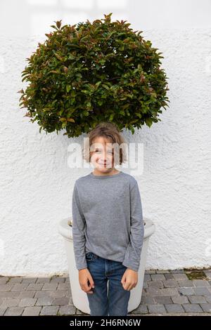 Portrait d'un jeune garçon devant un petit arbre contre un mur blanc Banque D'Images