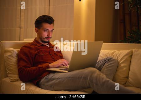 Un jeune homme avec une barbe en chemise rouge travaille avec un ordinateur portable et s'assoit sur le canapé le soir dans la maison. Banque D'Images