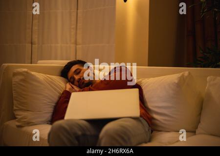 Un jeune homme avec une barbe en chemise rouge travaille avec un ordinateur portable et s'assoit sur le canapé le soir dans la maison. Banque D'Images