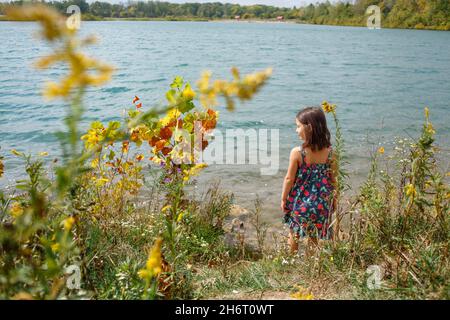 La petite fille se tient seule au bord de l'eau en regardant au-dessus de l'épaule Banque D'Images