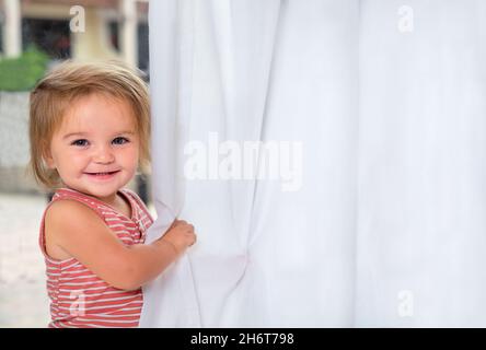Bébé mignon joue avec des rideaux.Portrait de l'enfant près de la fenêtre Banque D'Images