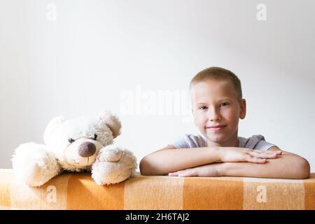 Le garçon joue avec un ours en peluche et regarde derrière le canapé.Joyeux enfant caucasien regardant la caméra sur l'arrière-plan d'un mur de lumière à la maison.Concept de loisirs enfant à la maison Banque D'Images