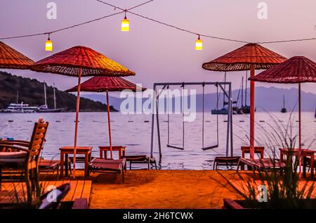 Parasols de plage, chaises longues et balançoires sous lanternes rouges sur la toile de fond de la mer, des îles, des montagnes et des yachts au coucher du soleil.Concept de vacances d'été Banque D'Images