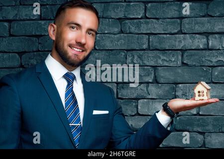 Homme en costume classique et cravate tient modèle maison en bois dans sa paume et sourires.Portrait d'un agent d'assurance ou d'un agent immobilier.Concept de vente et d'assurance de biens immobiliers.Arrière-plan avec espace de copie Banque D'Images