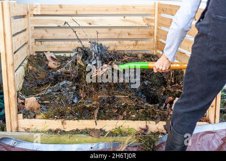 Un jardinier portant des bottes et des combinaisons en plastique tourne un tas de compost à l'aide d'une pelle ou d'une fourchette. Le travailleur transfère partiellement le matériau composté de Banque D'Images