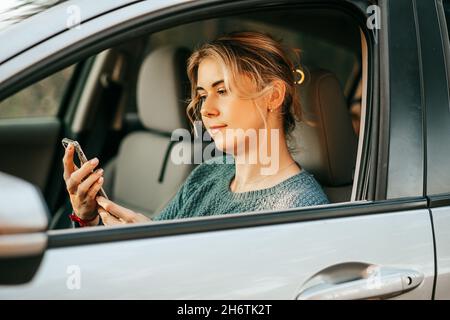 Photo de jeune belle femme se trouve derrière la roue de la voiture, regarde l'écran de téléphone mobile, la saisie sms massage utilise smartphone, système de navigation, carte, Banque D'Images