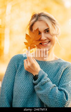 Portrait d'une jeune belle femme en pull tricoté de couleur bleue, caché derrière le feuillage d'automne, coquettishly regarde avec 1 œil dans la caméra, sourires, flir Banque D'Images