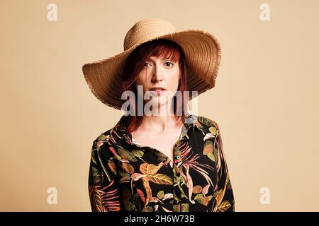 Jolie femme à tête rouge souriant avec une longue robe à fleurs et un chapeau.Portrait, avec un arrière-plan jaune. Banque D'Images