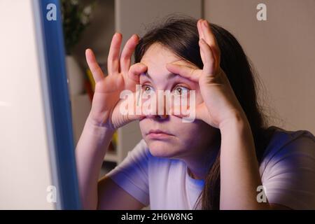 La femme se force à regarder dans l'écran de l'ordinateur, en ouvrant ses yeux larges avec ses mains Banque D'Images