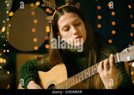 Une belle adolescente en bois de Noël et un chandail vert joue la guitare.Environnement domestique, arrière-plan sombre avec bokeh de la lumière de Noël Banque D'Images