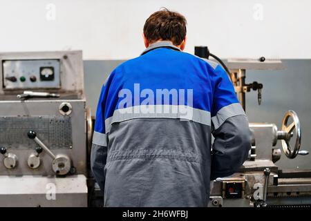 Turner se tient derrière le tour dans la salle de production et travaille.Vue de l'employé depuis l'arrière de la combinaison.Processus de travail authentique en production.Un vrai travailleur.Scène non stockée. Banque D'Images