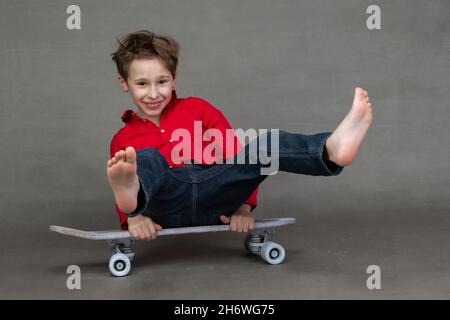 Drôle petit garçon assis sur un skateboard vintage pieds nus sur un fond gris. Banque D'Images