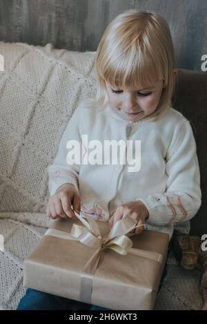 Une petite fille aux cheveux blonds ouvre un cadeau de Noël. Banque D'Images