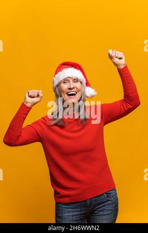 Portrait vertical d'une femme âgée hilarante à santa, dansant isolée sur une femme jaune d'âge moyen à cheveux gris célébrant le nouvel an Banque D'Images