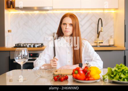 Portrait d'une jeune femme à tête rouge attrayante avec des yeux en colère tenant un gros couteau assis à une table avec planche à découper et regardant l'appareil photo Banque D'Images