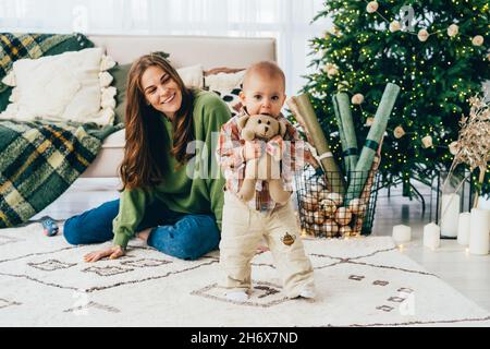 Une maman rieuse à tête rouge passe du temps avec son petit enfant joyeux dans la salle de séjour à l'heure de noël Banque D'Images